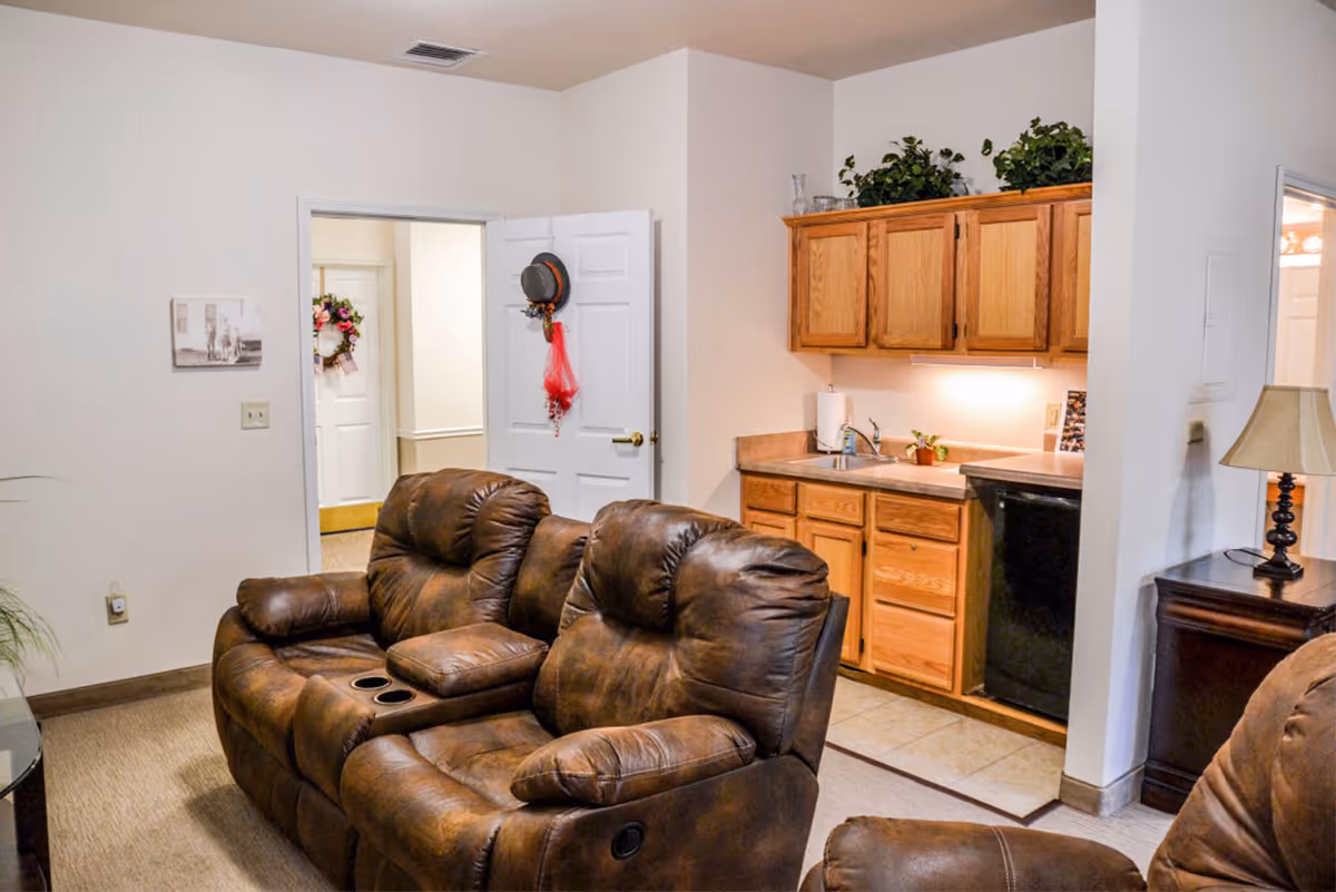 Interior view of a living area in The Bungalows at Springdale featuring two brown leather recliners with cup holders, a small kitchenette with wooden cabinets, a sink, and a black mini fridge. There is a white door with a hat and red scarf hanging on it, a lamp on a wooden side table, and some decorative plants on top of the cabinets.