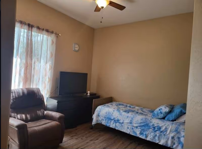 Small bedroom with a single bed in blue bedding, a recliner, a TV on a dresser, and a window with sheer curtains.