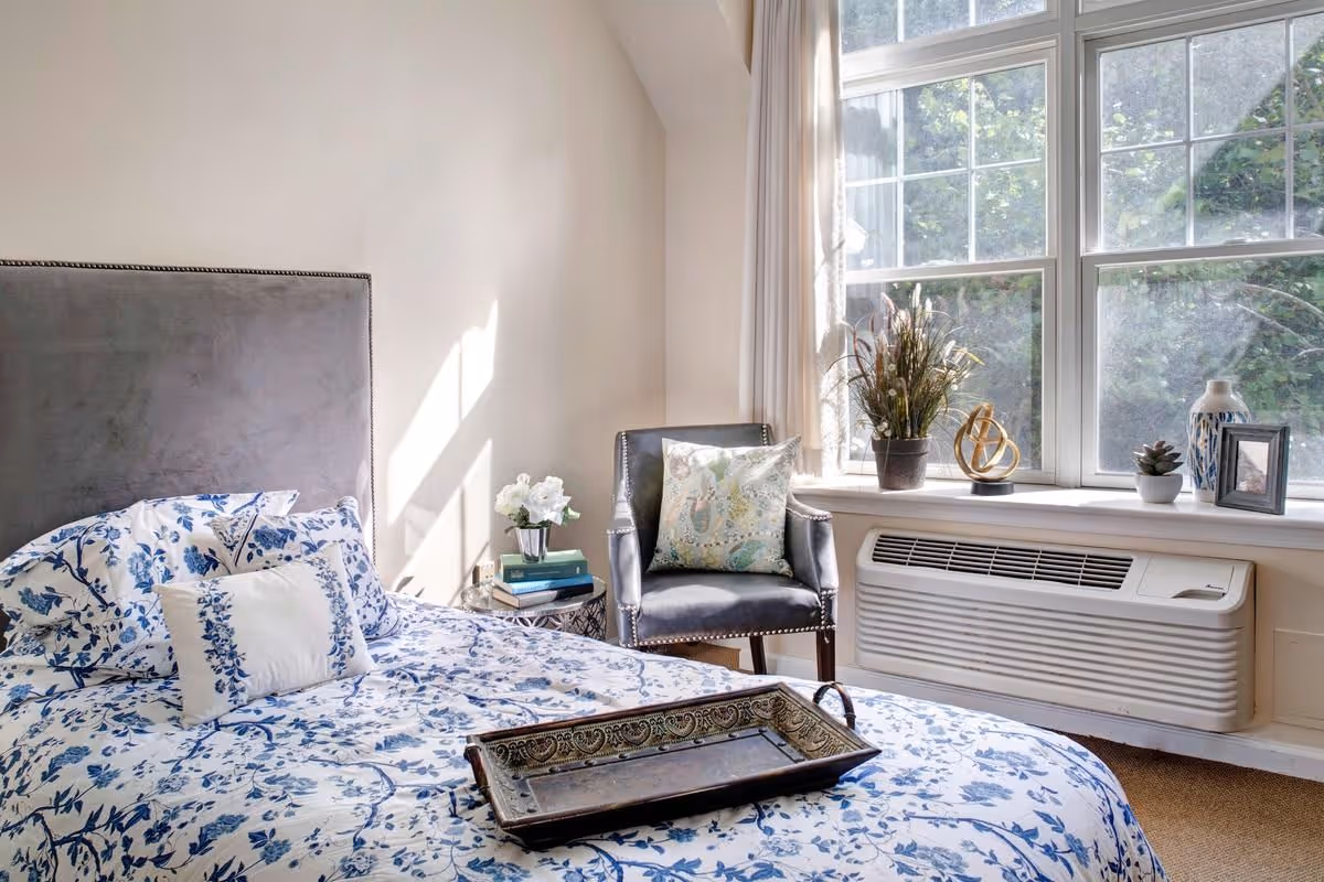 A bright bedroom with a bed covered in blue and white floral bedding, a gray upholstered headboard, and a decorative tray on the bed. Next to the bed is a small round side table with a vase of white flowers and stacked books. A gray armchair with a patterned cushion sits near a large window with beige curtains. The windowsill holds a potted plant, a decorative sculpture, a small succulent, a vase, and a framed photo. Below the window is a wall-mounted air conditioning unit.