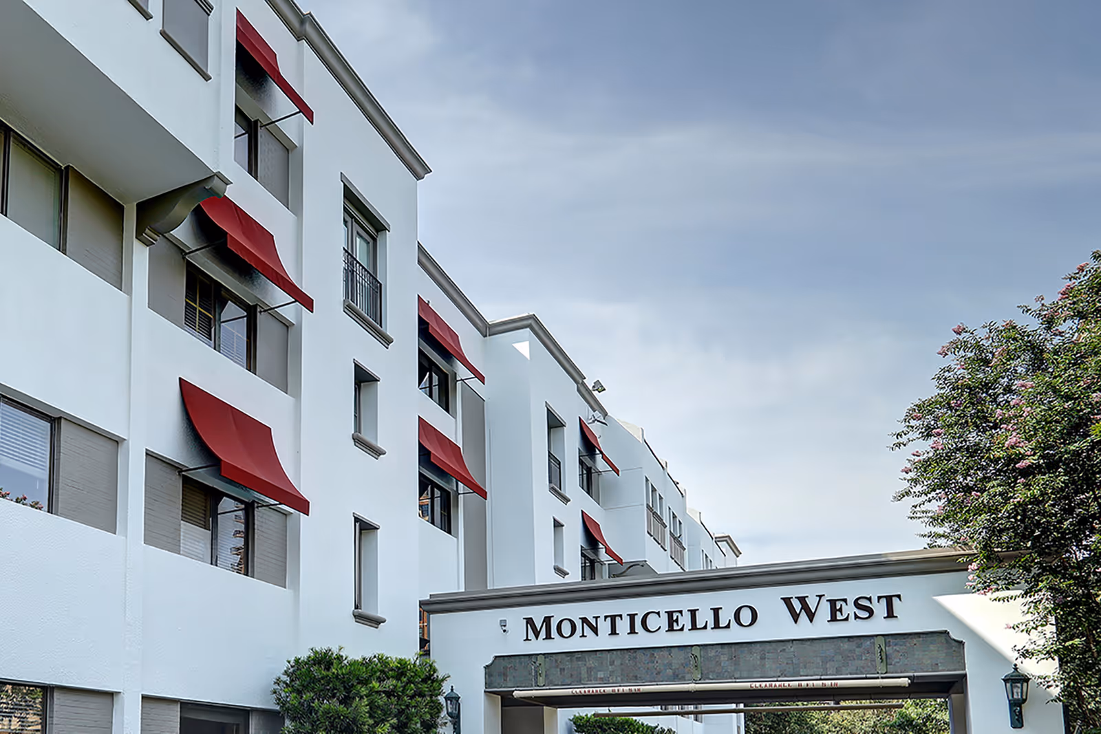 Exterior view of Monticello West building with white walls and red window awnings under a partly cloudy sky, with greenery and trees nearby.