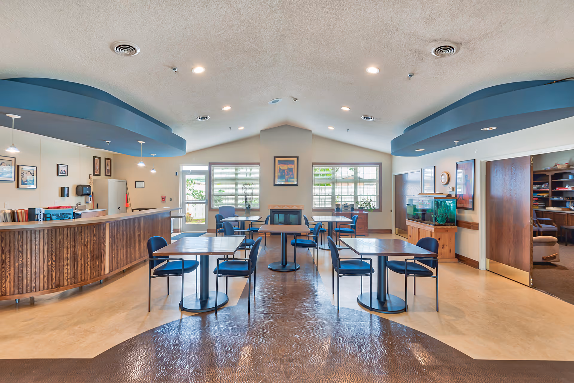 Bright communal dining room with several square tables and chairs, a long wooden service counter, large windows, and an aquarium.