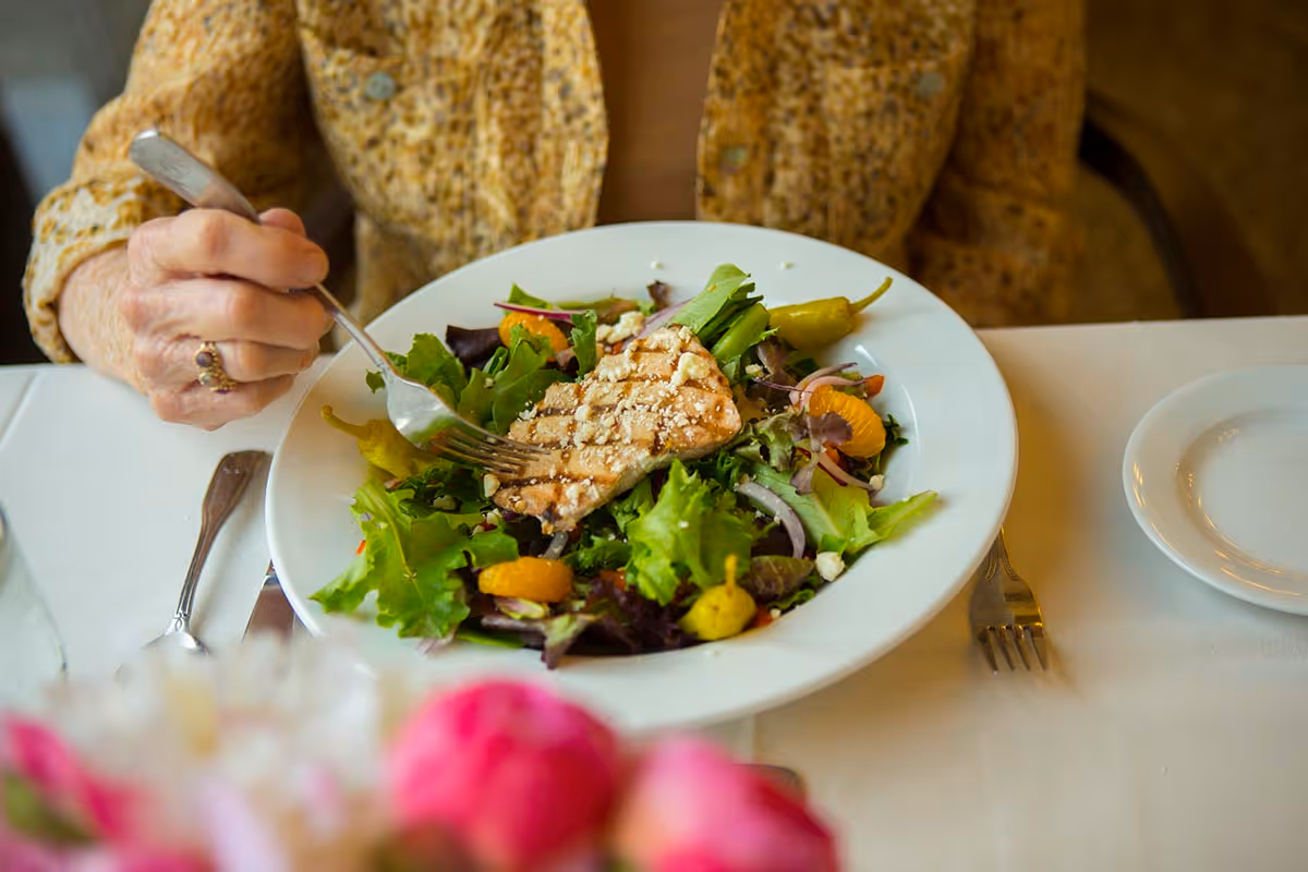 Person wearing a patterned jacket using a fork to eat a salad topped with grilled chicken at a set dining table.
