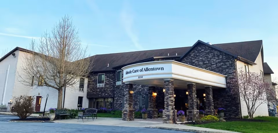 Exterior view of Abode Care of Allentown building with a covered entrance supported by stone pillars, a leafless tree and some flowering plants in front, under a clear blue sky.