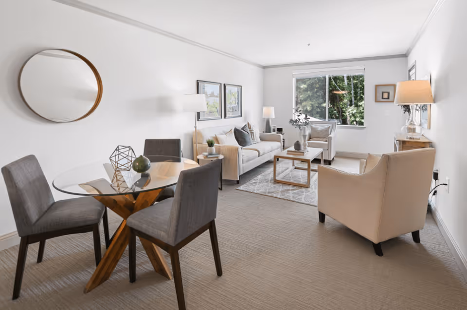 A bright and modern living room with a beige sofa, two armchairs, a glass coffee table on a patterned rug, and a round glass dining table with four gray upholstered chairs. The room has a large window showing green trees outside, white walls with framed artwork, and several lamps providing warm lighting.