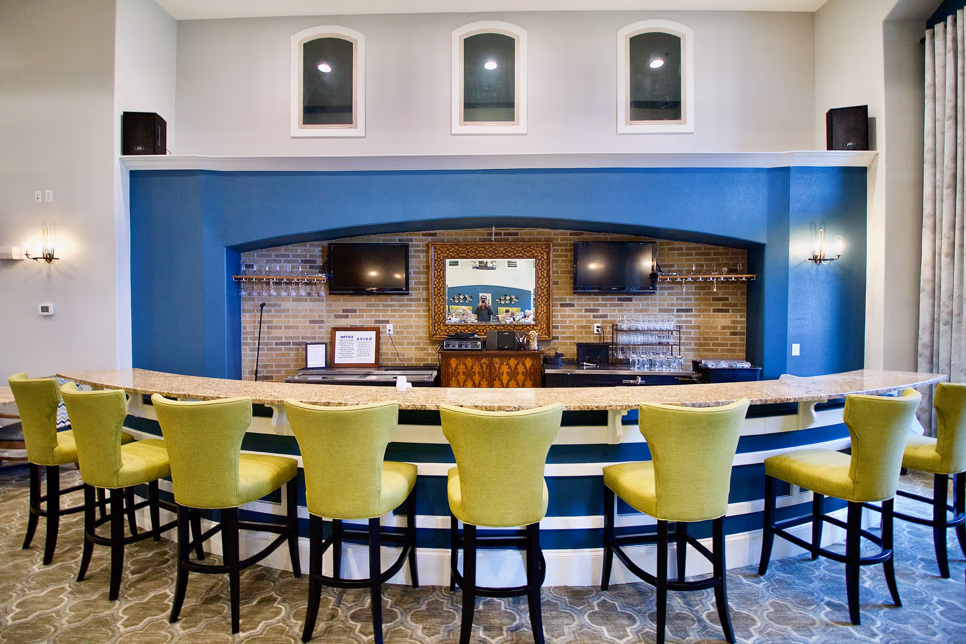 Interior view of a bar area with a curved granite countertop and eight green cushioned bar stools. The back wall features a blue accent with a brick inset, two mounted flat-screen TVs, a decorative mirror, and glassware on shelves. The floor has a patterned carpet, and the walls are painted light gray with sconces providing lighting.