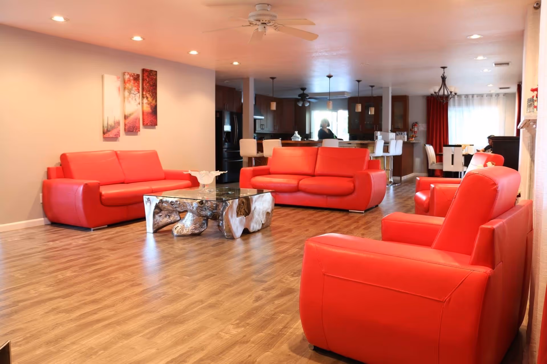 A spacious living room with bright red leather sofas and armchairs arranged around a unique wooden coffee table with a glass top. The room has wood flooring, recessed ceiling lights, and a ceiling fan. In the background, there is an open kitchen area with bar stools and a dining area with white chairs and red curtains.