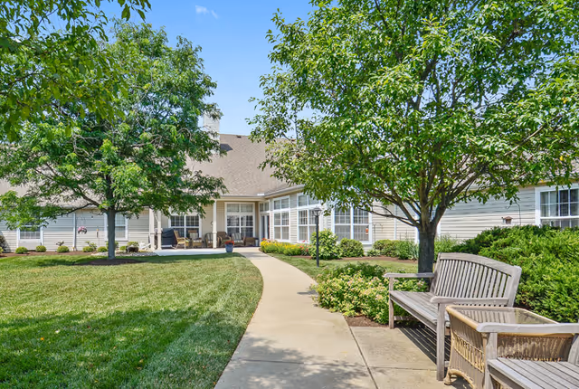 A sunny outdoor garden area at a senior living facility with a concrete pathway leading to a building entrance. The area features green grass, trees providing shade, and wooden benches along the path. The building has beige siding and large windows.