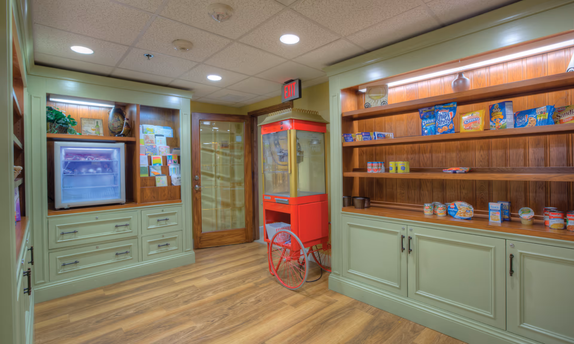 Well-lit interior snack area with built-in wooden shelves stocked with snacks, a small refrigerator, and a red popcorn machine on wood flooring.