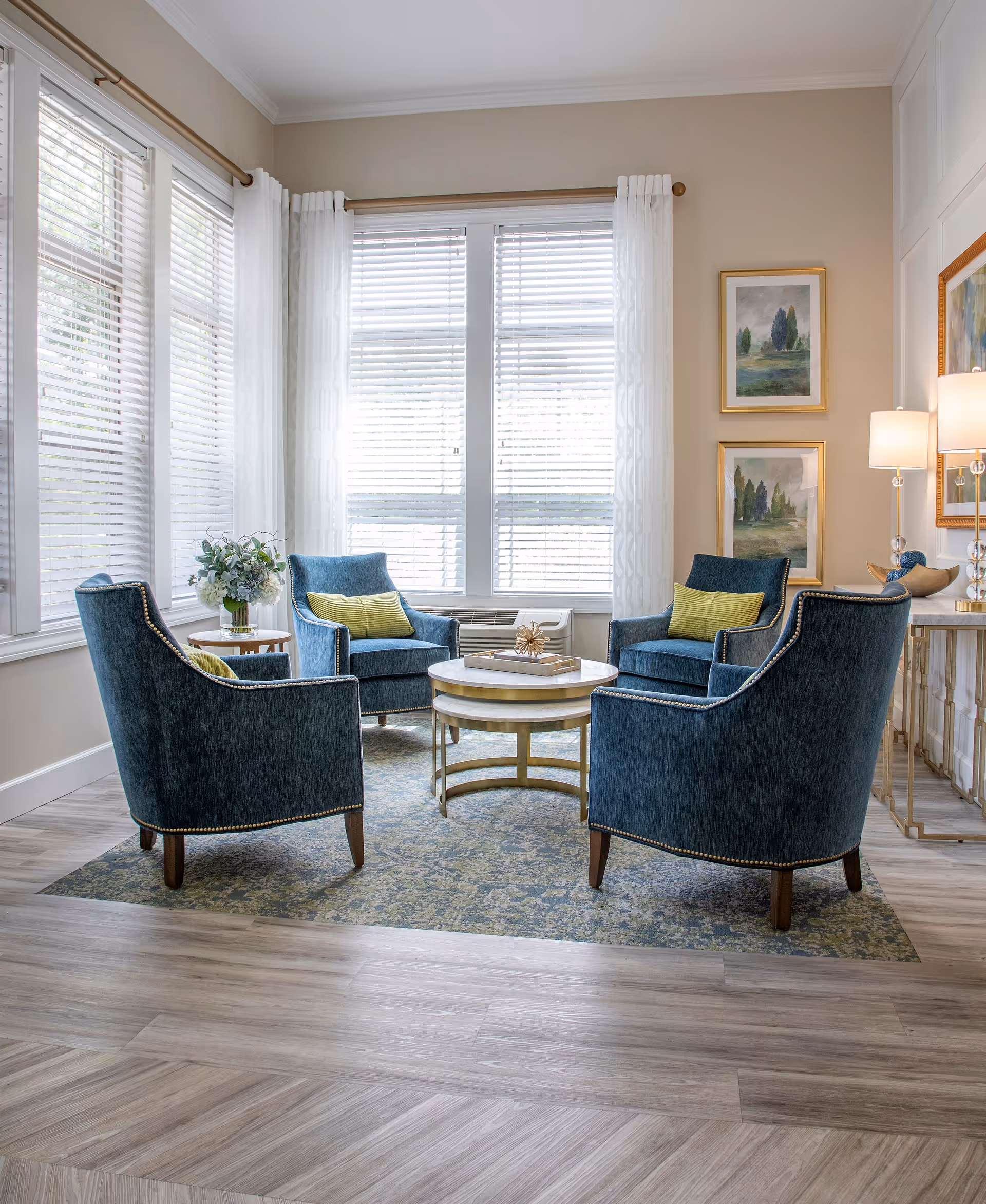 Sunlit sitting area with four blue upholstered armchairs arranged around a round coffee table in front of large windows.