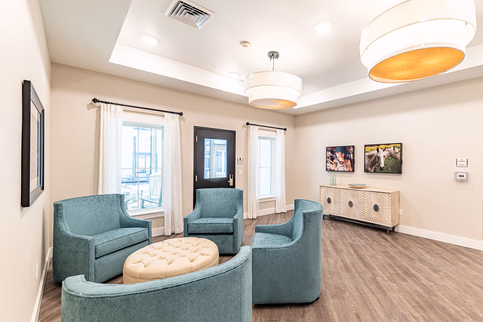 A bright and modern senior living facility common area with four teal upholstered chairs arranged around a beige tufted ottoman. The room has light-colored walls, two windows with white curtains, a black door with a window, wood flooring, and two large round ceiling lights. On the far wall, there is a decorative sideboard with two framed pictures above it, one of a woman and one of horses.