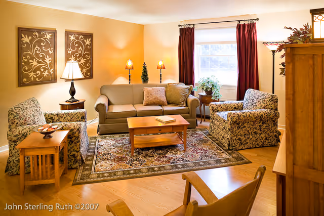 A cozy living room with a beige sofa, two floral-patterned armchairs, a wooden coffee table, and side tables. The room features warm yellow walls, a window with red curtains, decorative wall art, and several lamps providing soft lighting. A patterned area rug covers the wooden floor.