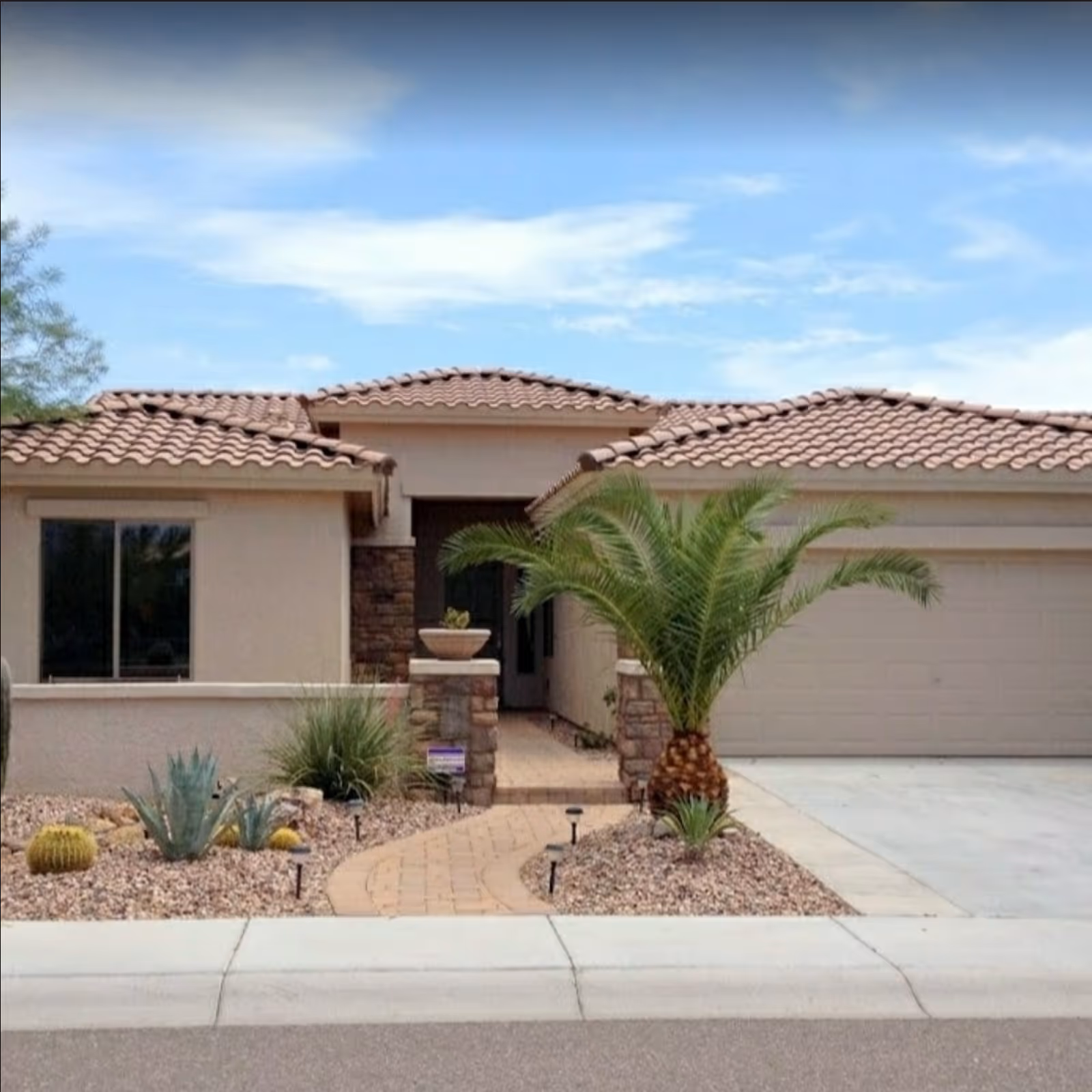 Front exterior view of a single-story house with a tiled roof, a paved walkway leading to the entrance, desert landscaping with various cacti and a palm tree, and a two-car garage.