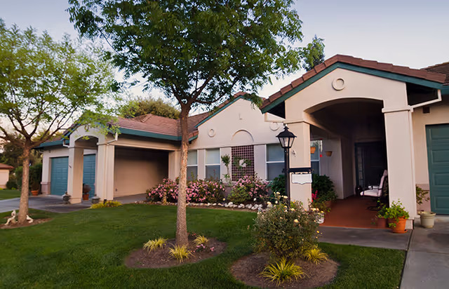 Exterior view of a single-story residential building with a covered porch, green garage doors, and well-maintained landscaping including trees, bushes, and flowers.