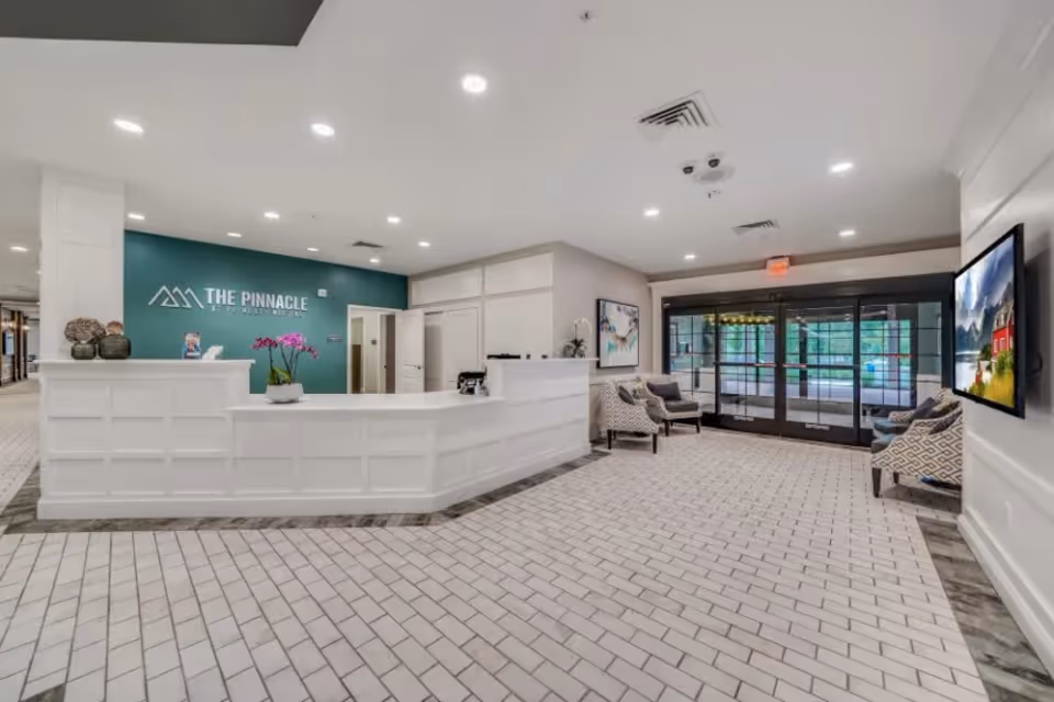 Reception area of The Pinnacle at Plymouth Meeting featuring a white front desk with decorative vases and a potted orchid, a teal accent wall with the facility's name and logo, seating area with patterned chairs, a large windowed entrance door, and a wall-mounted TV displaying a scenic image.
