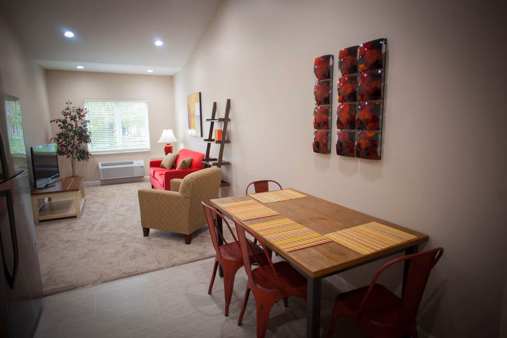 A cozy living and dining area with a wooden dining table set with four red metal chairs and four striped placemats. The living area features a red loveseat, a beige armchair, a floor lamp, a TV on a wooden stand, a potted plant, and a window with blinds. The walls are decorated with a modern art piece and a ladder-style shelf.