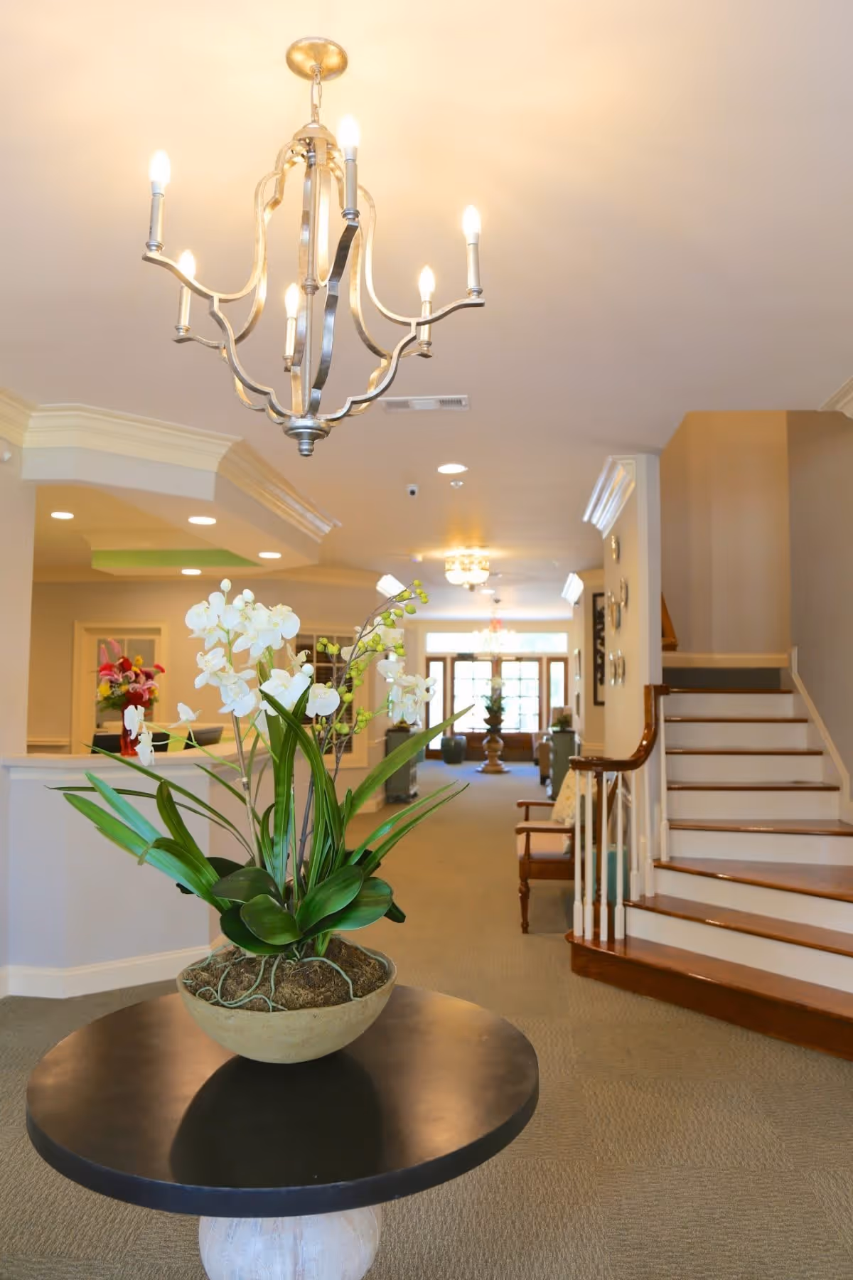 Interior view of a senior care facility lobby featuring a round table with a large potted white orchid plant in the foreground, a chandelier hanging from the ceiling, a staircase with wooden handrails on the right, and a hallway leading to seating areas and large windows in the background.