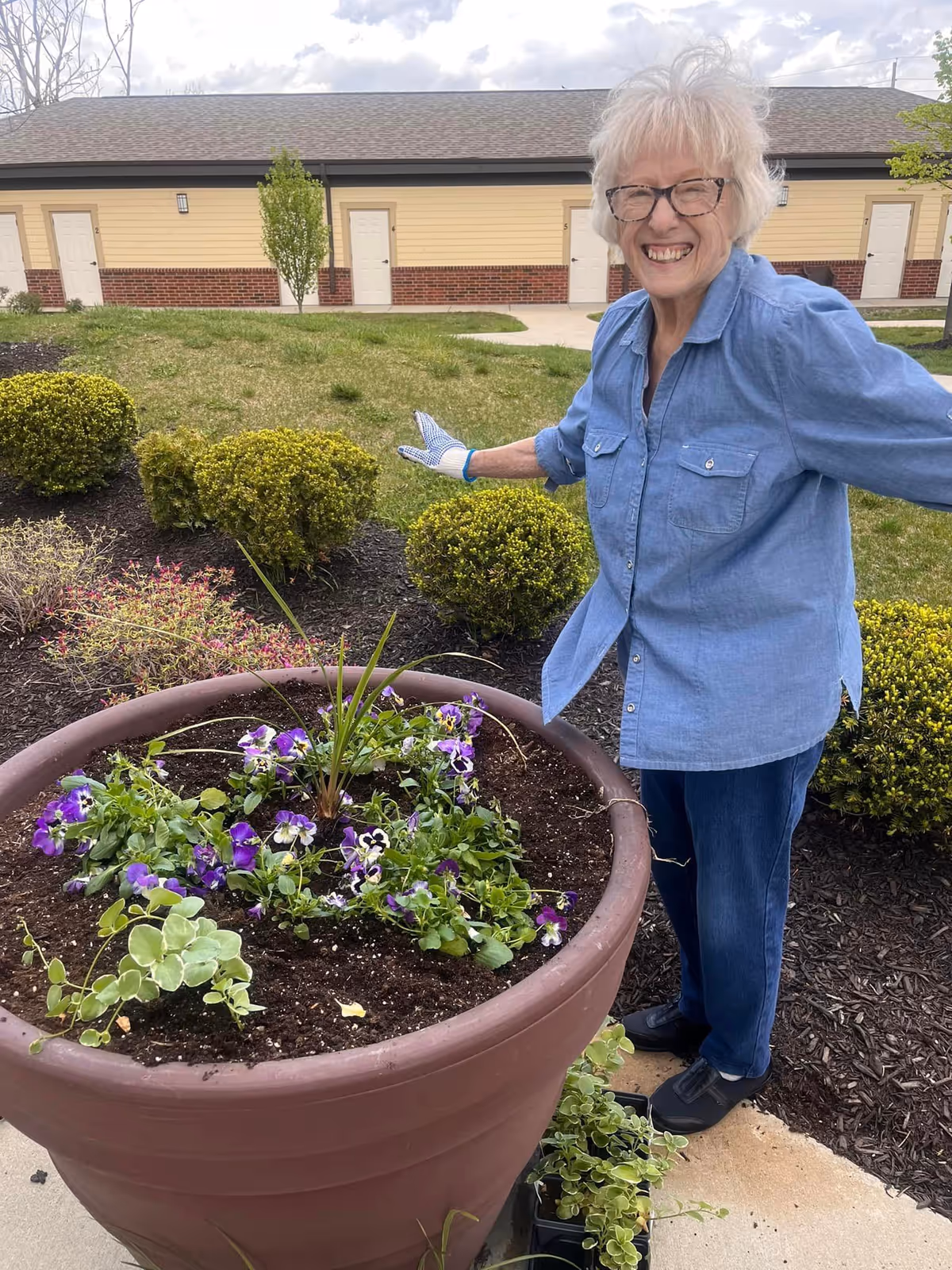 An elderly woman wearing glasses, a blue shirt, jeans, and gardening gloves is smiling and standing next to a large planter filled with soil and purple flowers. Behind her is a grassy area with trimmed bushes and a building with multiple doors.