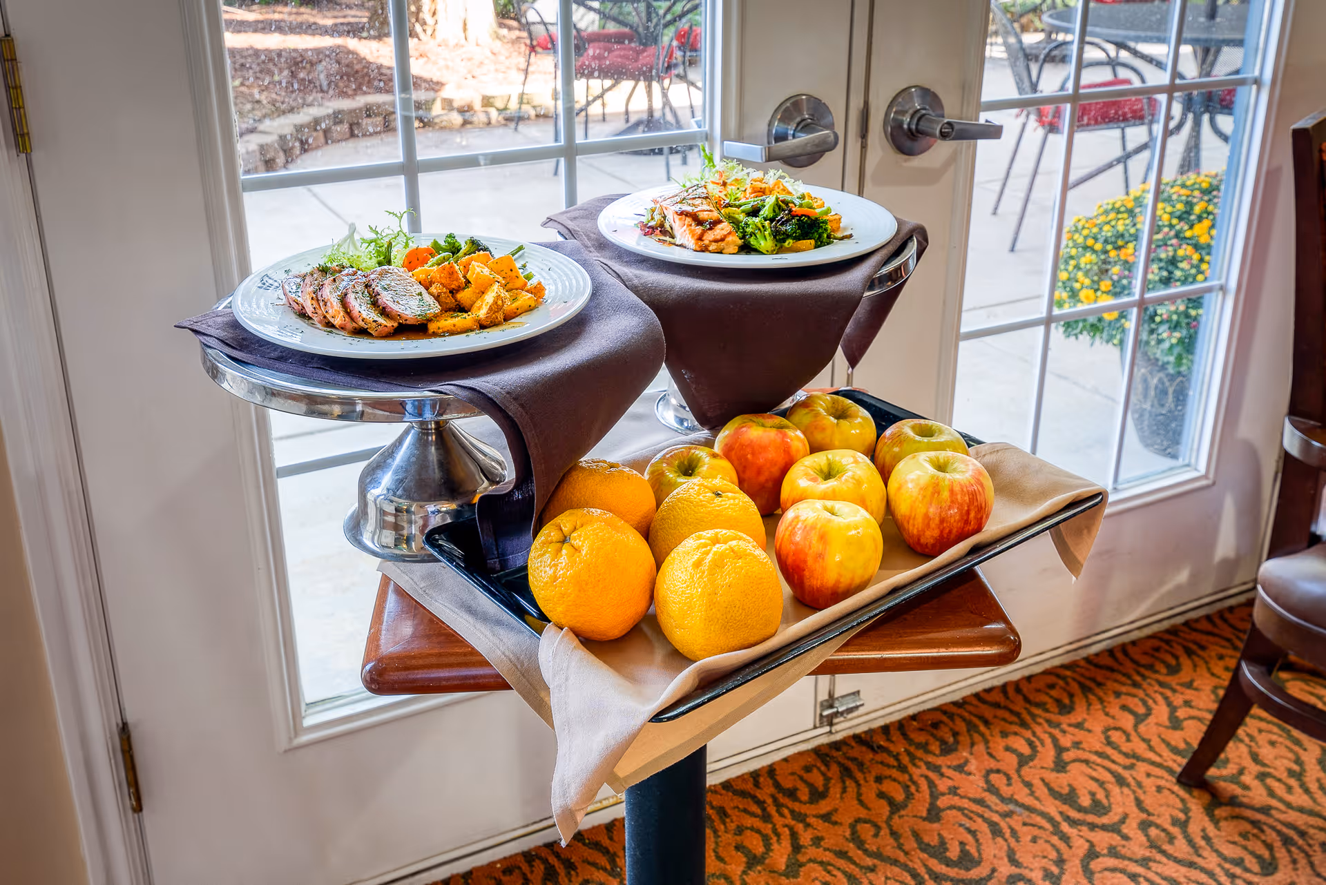 A serving cart holding two plated meals and a tray of apples and oranges placed in front of glass double doors.