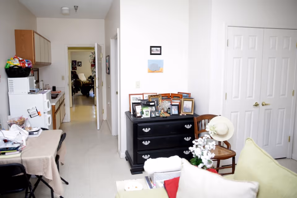 Interior view of a senior living facility room showing a small kitchenette area with a microwave, cabinets, and a basket of snacks on top of the microwave. A table with a beige tablecloth and black folding chairs is on the left. In the foreground, there is a black dresser with multiple framed photos on top, a wooden chair with a white hat hanging on it, and a green couch with pillows. The room has white walls and a hallway leading to another room with more furniture visible.