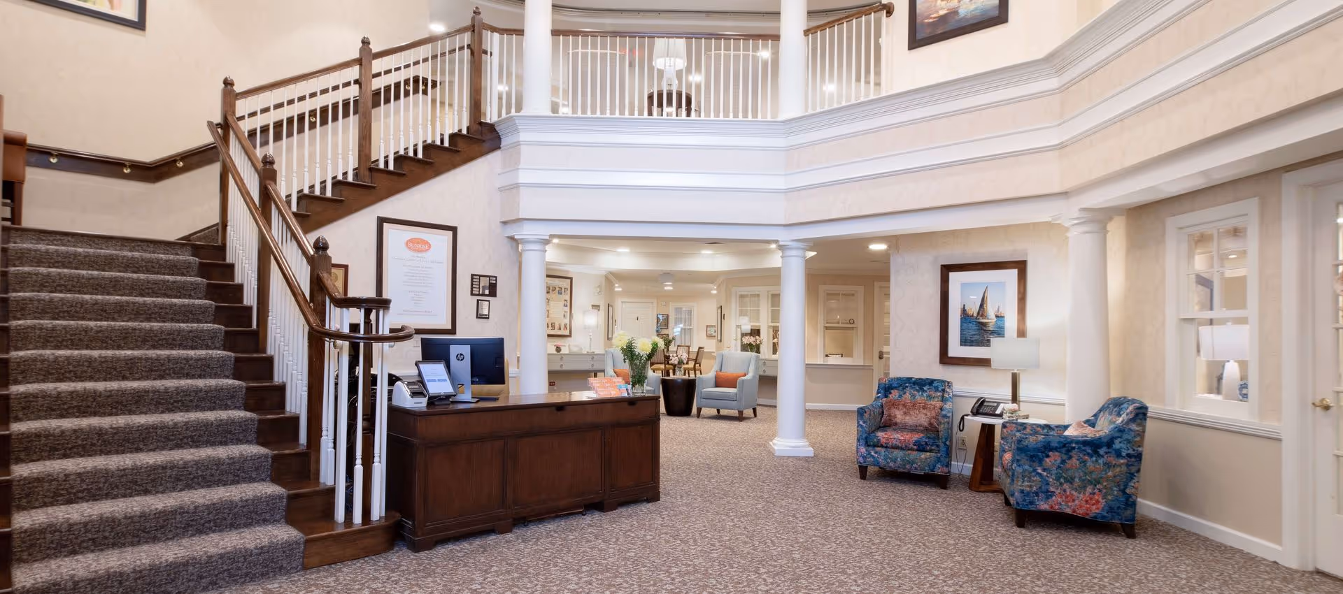 Interior view of a senior living facility lobby with a carpeted staircase on the left, a wooden reception desk with computer monitors, and seating area with floral upholstered chairs and a side table with a lamp and phone. The space features cream-colored walls, white columns, framed artwork, and a second-floor balcony with white railings.