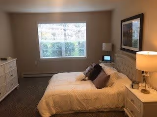 A neatly made bed with white bedding and several pillows in a softly lit bedroom. There is a nightstand with a lamp and a small digital clock on one side of the bed, and a white dresser on the opposite side. A large window with blinds lets in natural light, and a framed picture hangs on the wall above the bed.