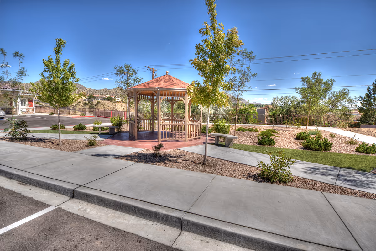 Outdoor area with a wooden gazebo surrounded by small trees, shrubs, and landscaped gravel. There are concrete sidewalks and benches near the gazebo, with a clear blue sky and hills in the background.