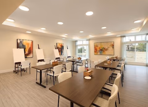 A bright, spacious activity or meeting room with several tables and chairs arranged neatly. There are four easels with blank canvases along one wall, and colorful abstract paintings hang on the walls. Large windows let in natural light, and the floor has light-colored wood or laminate flooring.