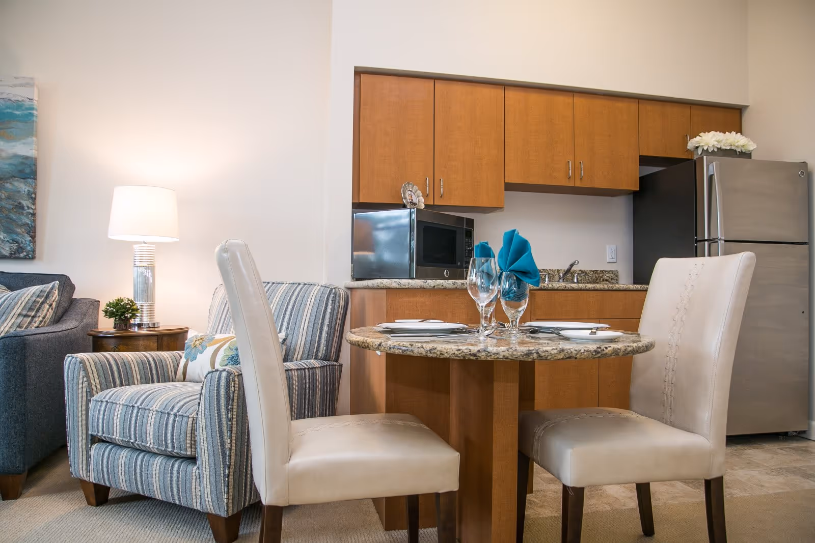 A small dining area with a round granite table set for two with plates, silverware, wine glasses, and blue cloth napkins. Behind the table is a kitchenette with wooden cabinets, a microwave, a sink, and a stainless steel refrigerator. To the left, there is a striped armchair with a floral cushion and a side table with a lamp and a small plant. Part of a blue sofa and a wall painting are also visible.