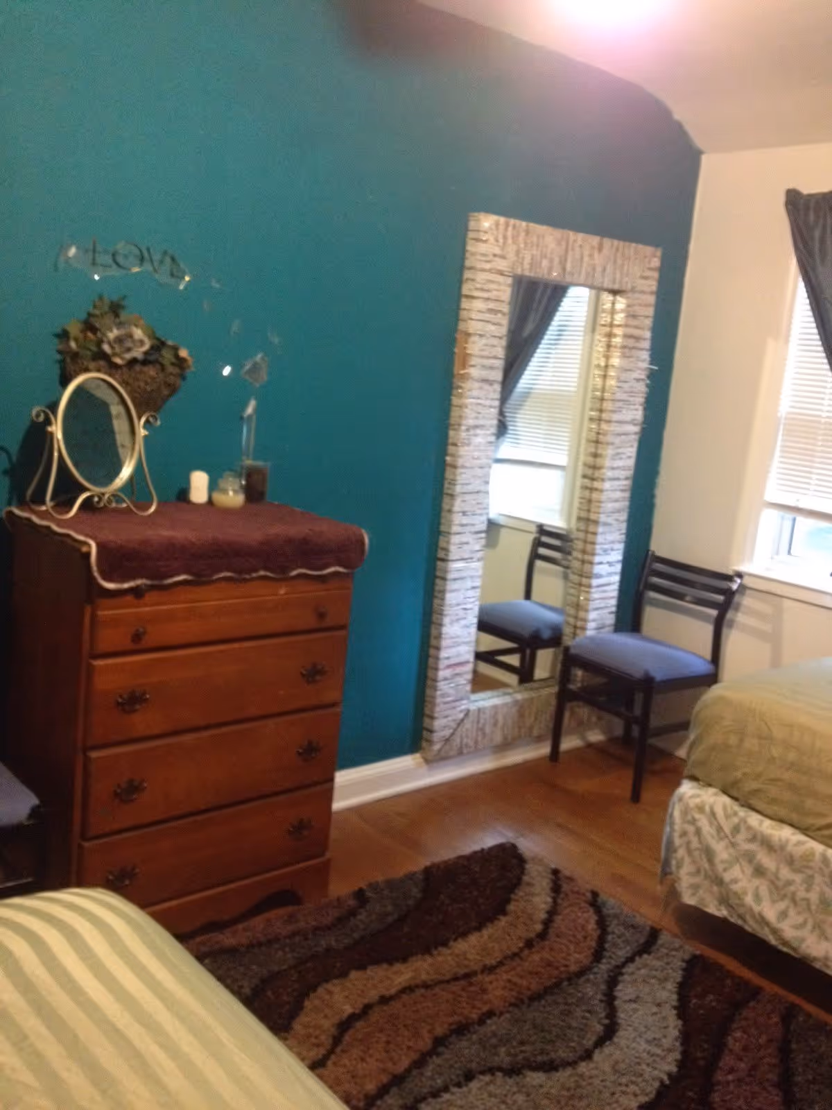 Cozy bedroom with a teal accent wall, wooden dresser, large floor mirror, chairs, and beds.