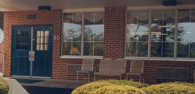 Brick building front showing double teal entrance doors, large windows, metal chairs, and shrubs.