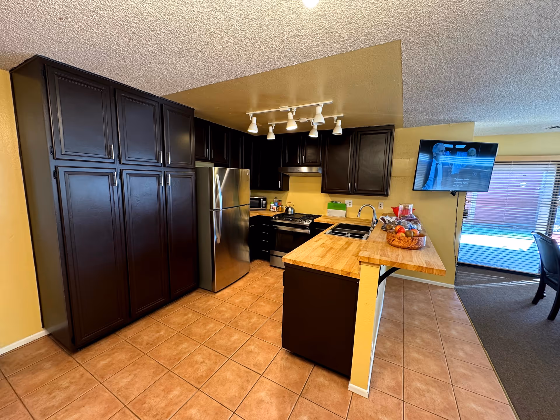 A kitchen area with dark brown cabinets, a stainless steel refrigerator, stove, and microwave. The countertops are wooden, and there is a sink with a faucet. On the counter, there is a bowl of fruit and some other items. The floor is tiled, and a TV is mounted on the wall near a window with blinds. The kitchen opens into a living area with carpeted flooring.