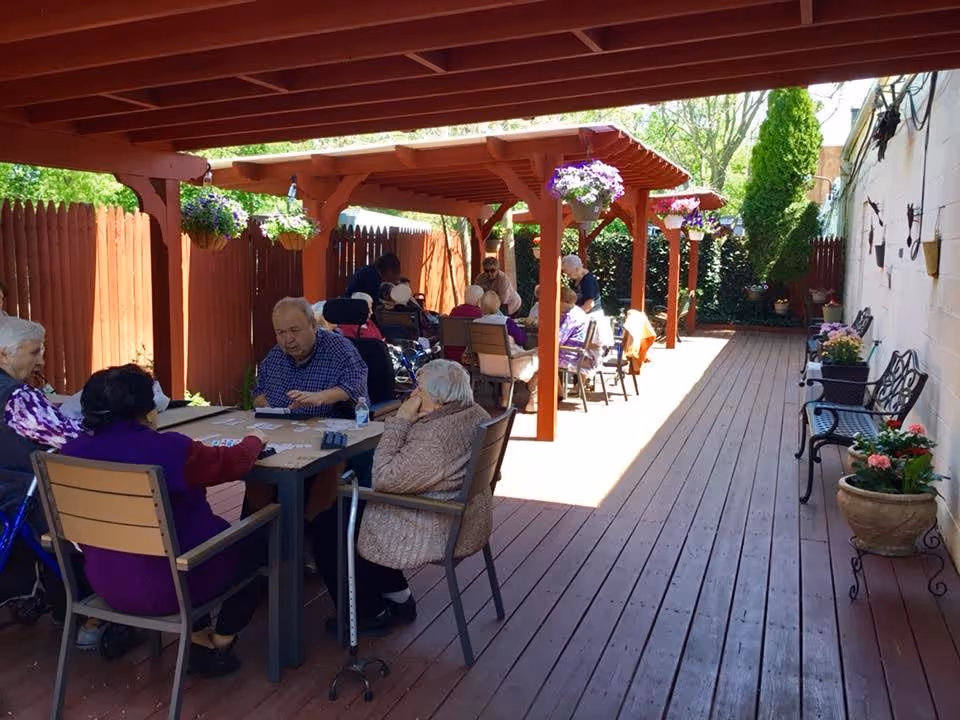 A group of elderly people sitting at tables under a wooden pergola on a wooden deck, playing cards and socializing. The area is fenced with a red wooden fence and decorated with hanging flower pots and potted plants. There are benches along a white wall on the right side.