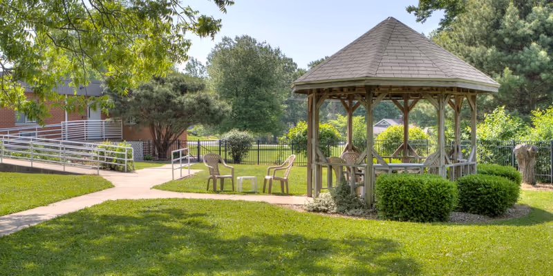 Outdoor garden area at Carlyle Healthcare Center featuring a wooden gazebo with a shingled roof, surrounded by green bushes and trees. There are plastic chairs and a small table on the paved walkway near the gazebo. The background shows a brick building and a metal fence enclosing the area.