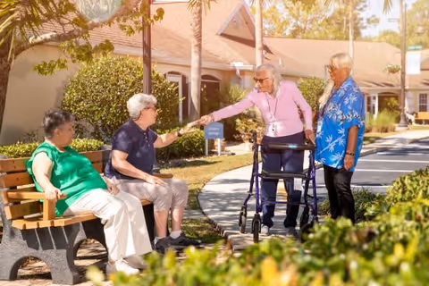 Four elderly individuals outdoors at Palm Cottages of Rockledge. Two women are sitting on a bench, one wearing a green shirt and white pants, the other wearing a navy blue shirt and light-colored pants. Another woman with a walker, dressed in a pink top and dark pants, is reaching out to shake hands with the woman in the navy blue shirt. A man in a blue Hawaiian shirt and dark pants stands nearby. The background shows a sunny day with trees, bushes, and a building with a beige exterior.