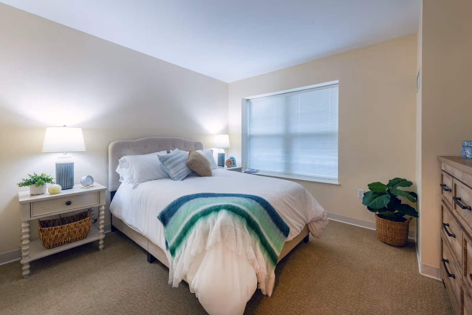 A cozy bedroom with a neatly made bed featuring white linens, a green and blue striped throw blanket, and several pillows. There are two bedside tables with lamps and decorative items, a large window with closed blinds, a wooden dresser, and a potted plant in the corner.