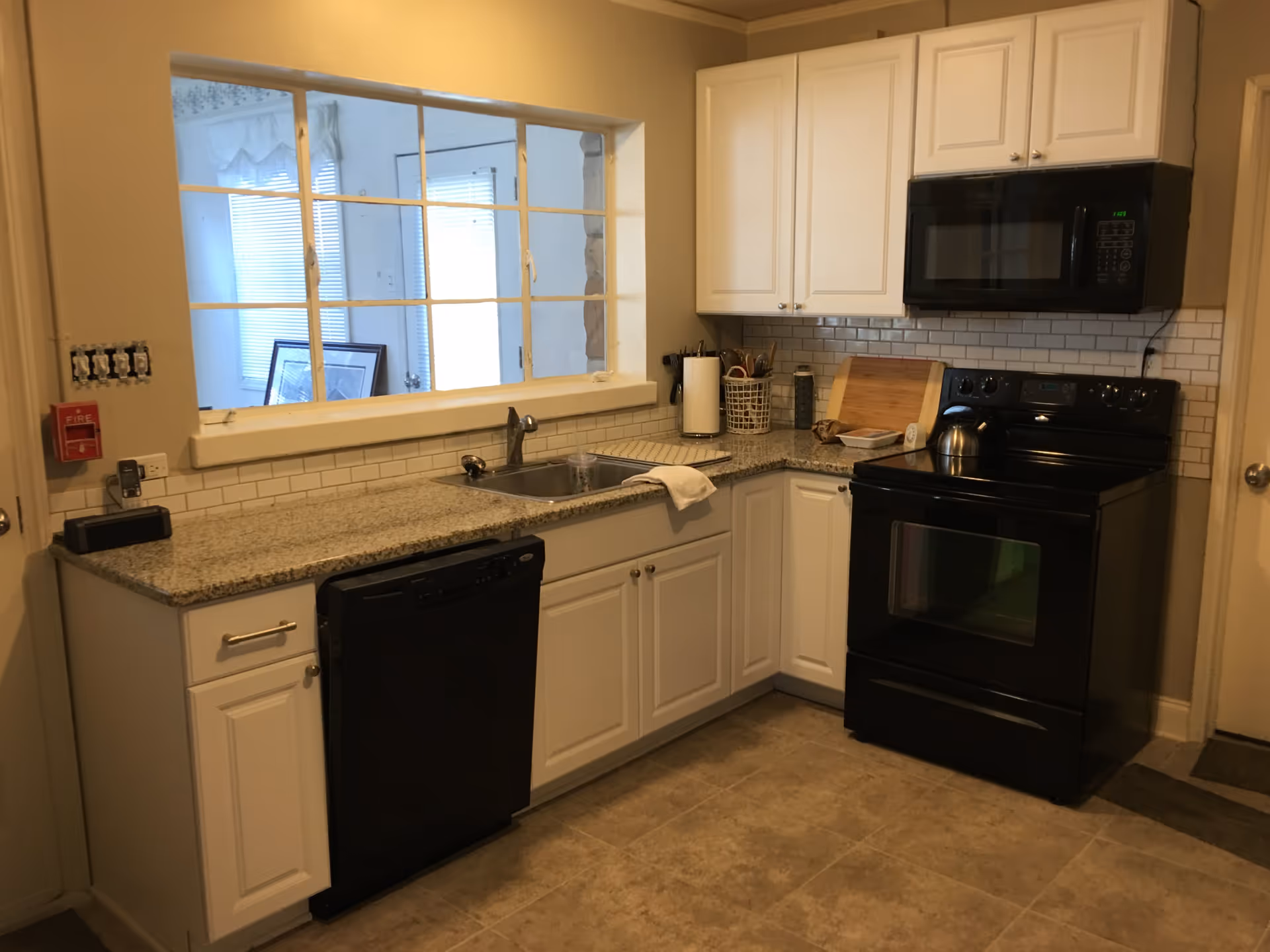 A kitchen with white cabinets, granite countertops, a black dishwasher, black stove with oven, and a black microwave above the stove. There is a stainless steel sink under a large window with multiple panes looking into another room. The backsplash is white subway tile, and the floor is tiled in a beige color. Various kitchen utensils and a paper towel holder are on the countertop.
