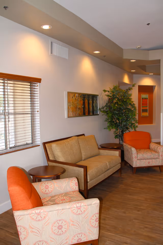 A cozy living room area with a beige sofa and two patterned armchairs with orange cushions. There are two round wooden side tables, a large green potted plant, and a window with wooden blinds. The walls are light-colored with recessed lighting and a colorful abstract painting hanging above the sofa.