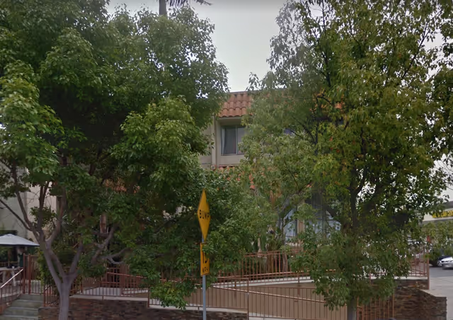 Exterior view of a senior living facility entrance obscured by trees and a yellow street sign.