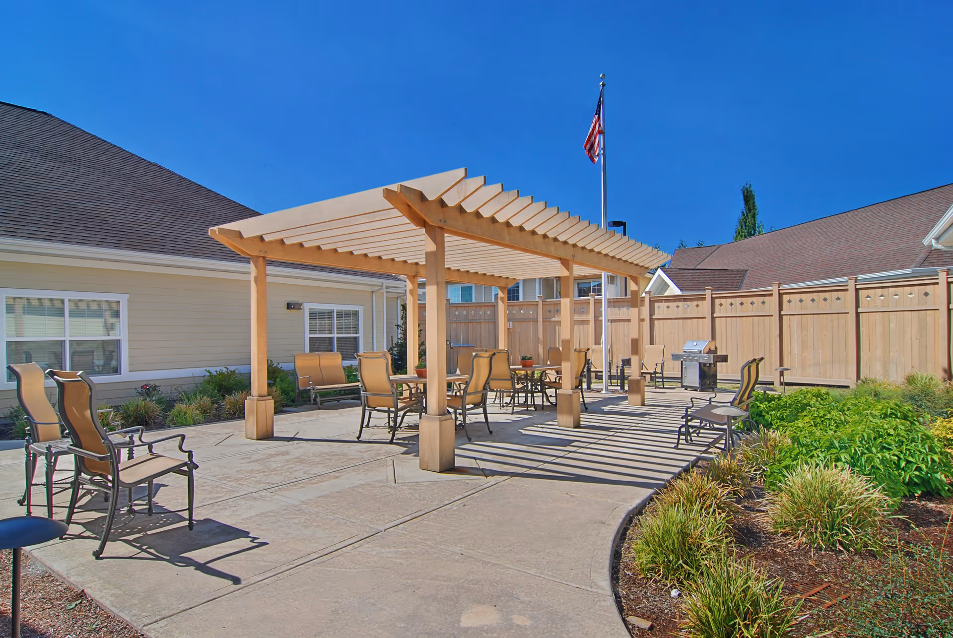 Sunny outdoor courtyard with a wooden pergola, patio tables and chairs, a grill, and an American flag near residential buildings.