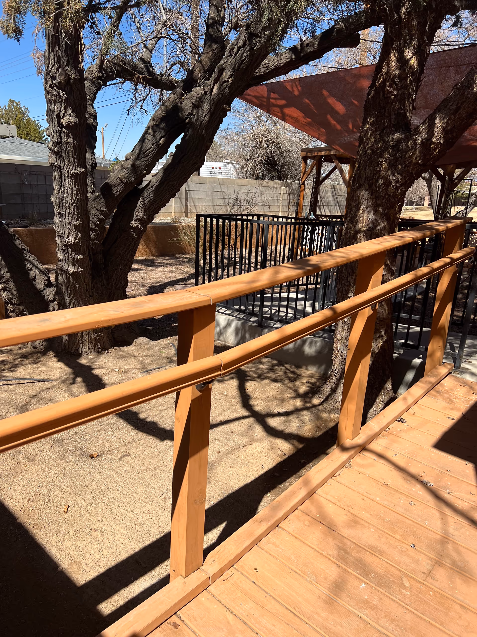 Outdoor area with a wooden ramp railing, large trees casting shadows, and a fenced section under a red shade canopy. The ground is covered with dirt and there is a concrete block wall in the background.