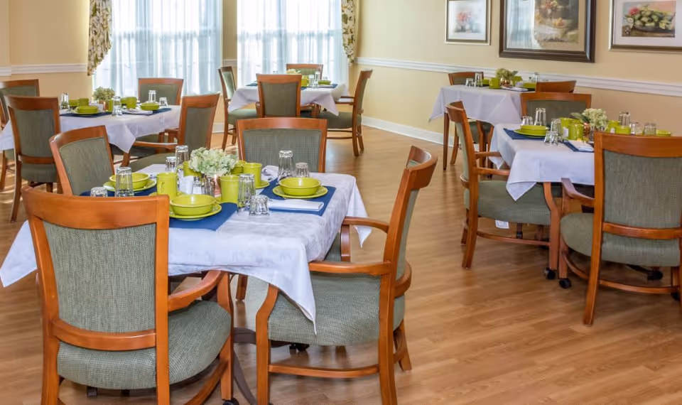 A bright dining room with several tables covered in white tablecloths, each set with green plates, cups, and silverware. Wooden chairs with green upholstery surround the tables. The room has light-colored walls, framed artwork, and large windows with sheer curtains allowing natural light to fill the space.