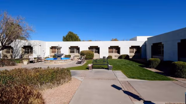 Outdoor courtyard area of a senior living facility with a central water fountain surrounded by benches and chairs, green grass, bushes, and a clear blue sky.