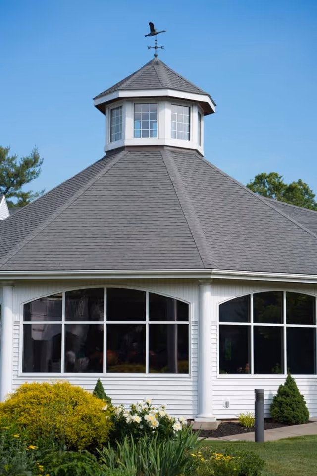 Exterior view of a white building with a gray shingled octagonal roof topped with a small cupola and a weather vane. The building has large arched windows and is surrounded by green bushes, flowering plants, and a well-maintained lawn under a clear blue sky.