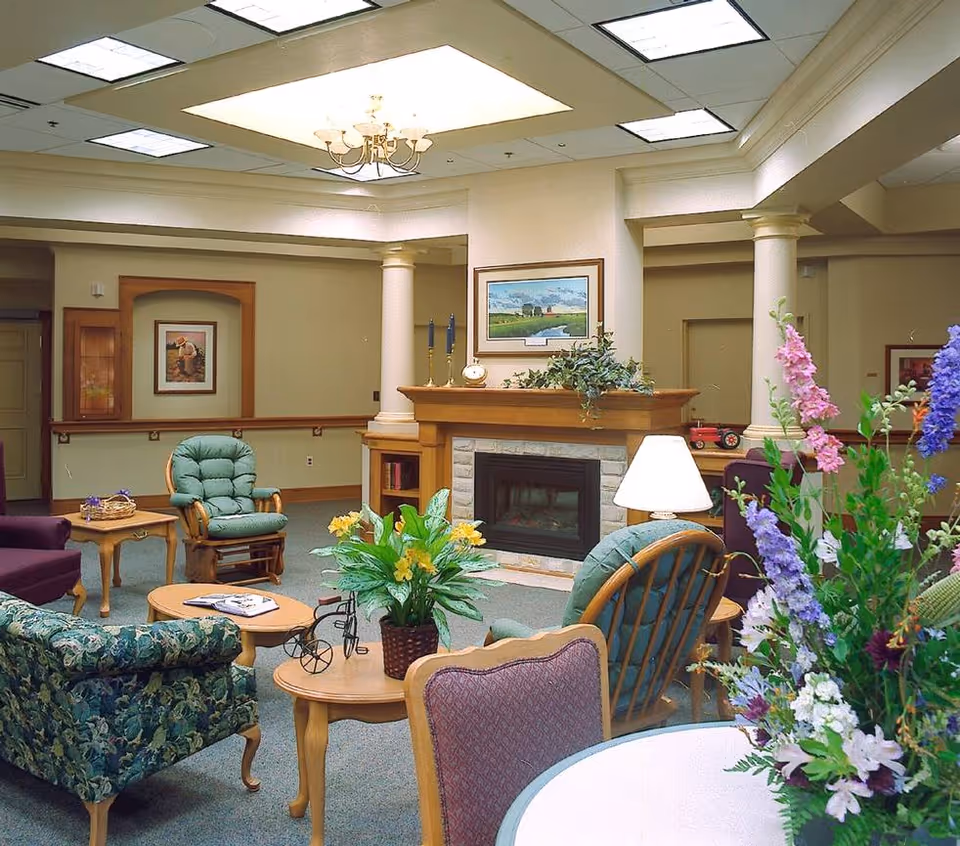 A cozy living room area in a senior living facility featuring a fireplace with a wooden mantle, surrounded by cushioned chairs and small wooden tables. The room has decorative plants and flowers, a chandelier, and framed artwork on the walls. The space is warmly lit and inviting.