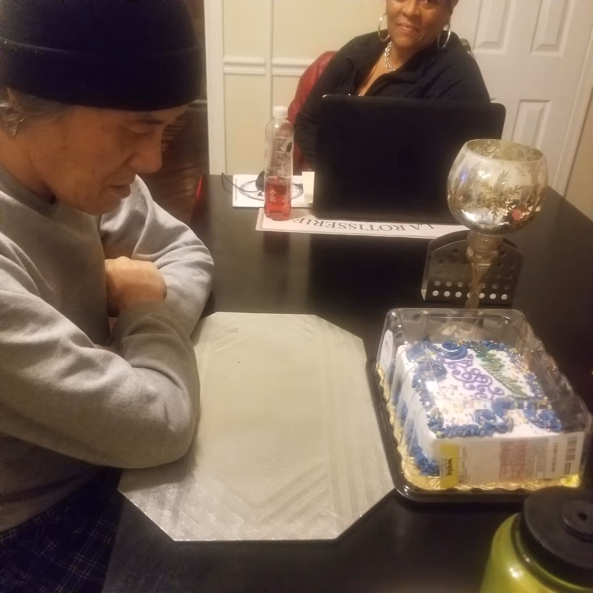 Two people sitting at a dark dining table. One person is looking down at the table with arms crossed, wearing a black beanie and gray sweatshirt. The other person is sitting across the table with a laptop in front of them, wearing hoop earrings and a black top. On the table, there is a decorated cake in a plastic container, a decorative candle holder, a bottle of drink, and some papers.