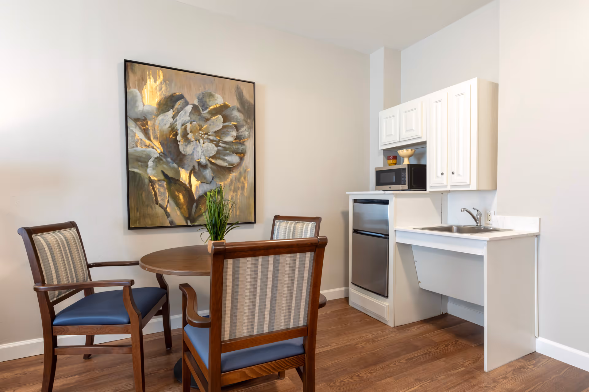 A small dining area with a round wooden table and three wooden chairs with blue cushions and striped backs. On the table is a small green plant. Behind the table is a large framed painting of a flower with gold accents. To the right is a kitchenette area with white cabinets, a stainless steel mini fridge, a microwave, and a sink with a faucet. The floor is wooden and the walls are painted light beige.