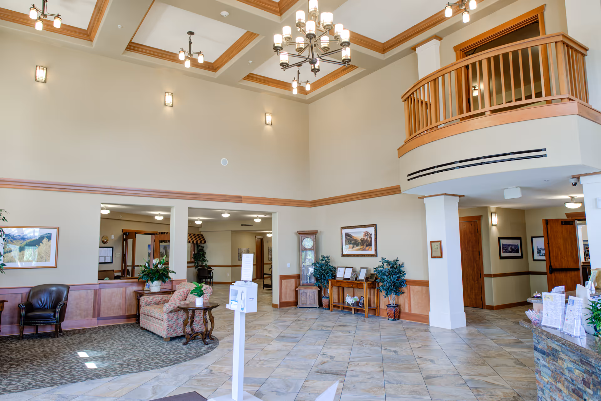 Spacious senior living facility lobby with seating, plants, high ceilings, chandeliers and a second-floor balcony.