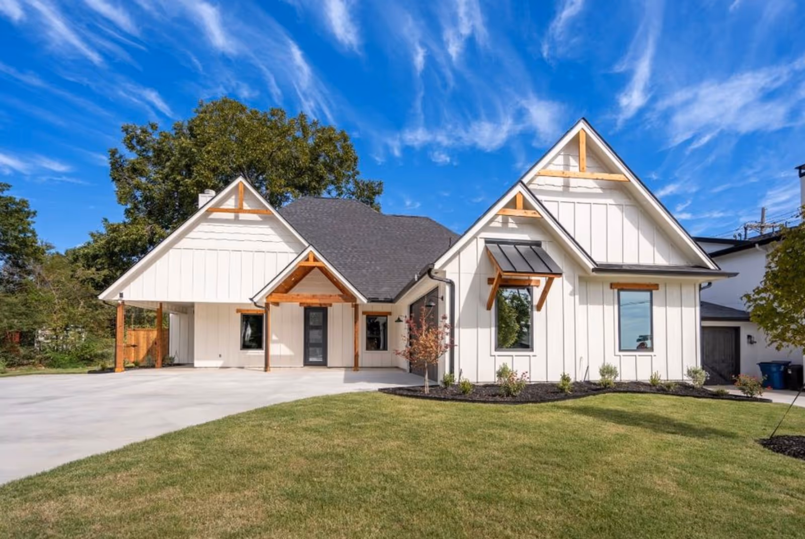 A modern white house with a steep roof and wooden accents, surrounded by a well-maintained lawn and a concrete driveway under a bright blue sky with wispy clouds.