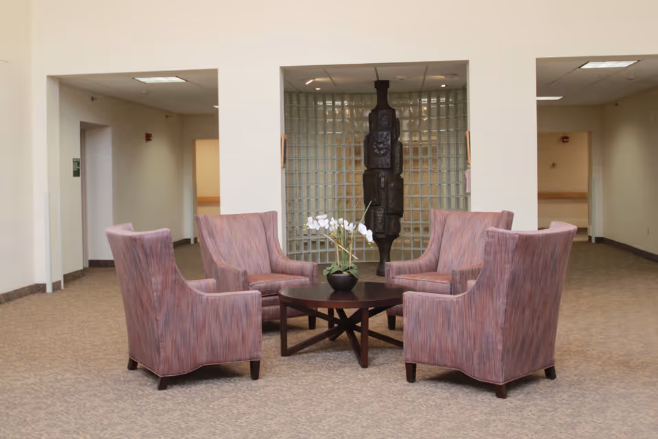 A seating area in a senior living facility with four upholstered armchairs arranged around a round wooden coffee table holding a small potted orchid. The background features a glass block wall with a dark sculptural art piece in front of it. The space has beige walls and carpeted flooring.