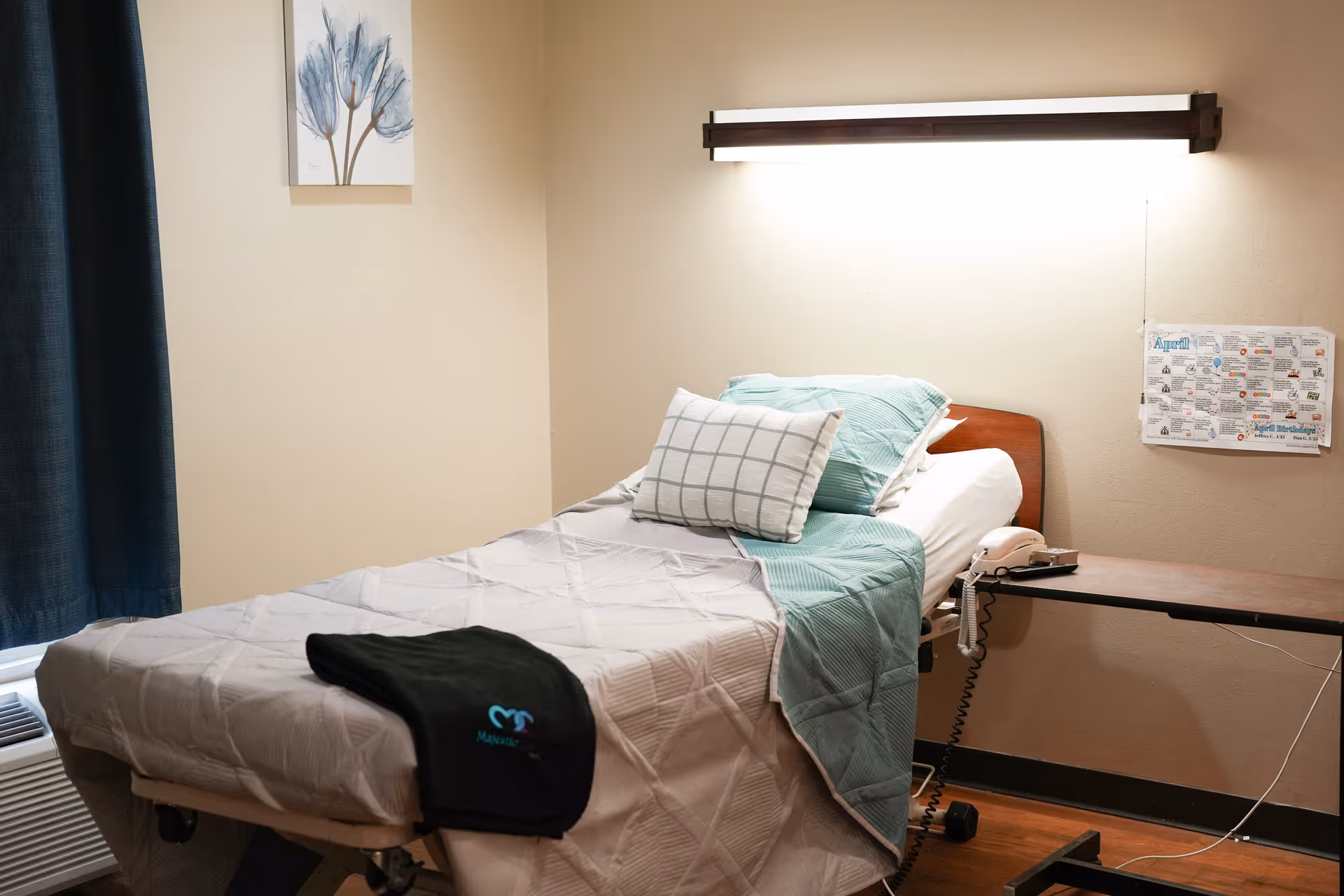 A neatly made single bed in a room with light beige walls, featuring a white and green checkered pillow, a green blanket, and a black cloth with the Majestic Care logo on it. There is a wall-mounted light above the bed, a small table with a telephone beside the bed, a window with dark curtains, and a framed floral artwork on the wall.